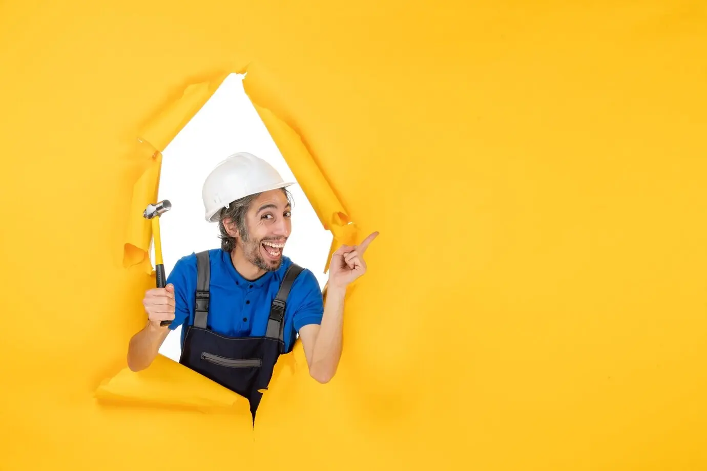 Vorderansicht eines männlichen Bauarbeiters in Uniform mit Hammer vor einer gelben Wand; Arbeiter, Mann, Bauunternehmer, Architektur, Farbe, Gebäude, Job.