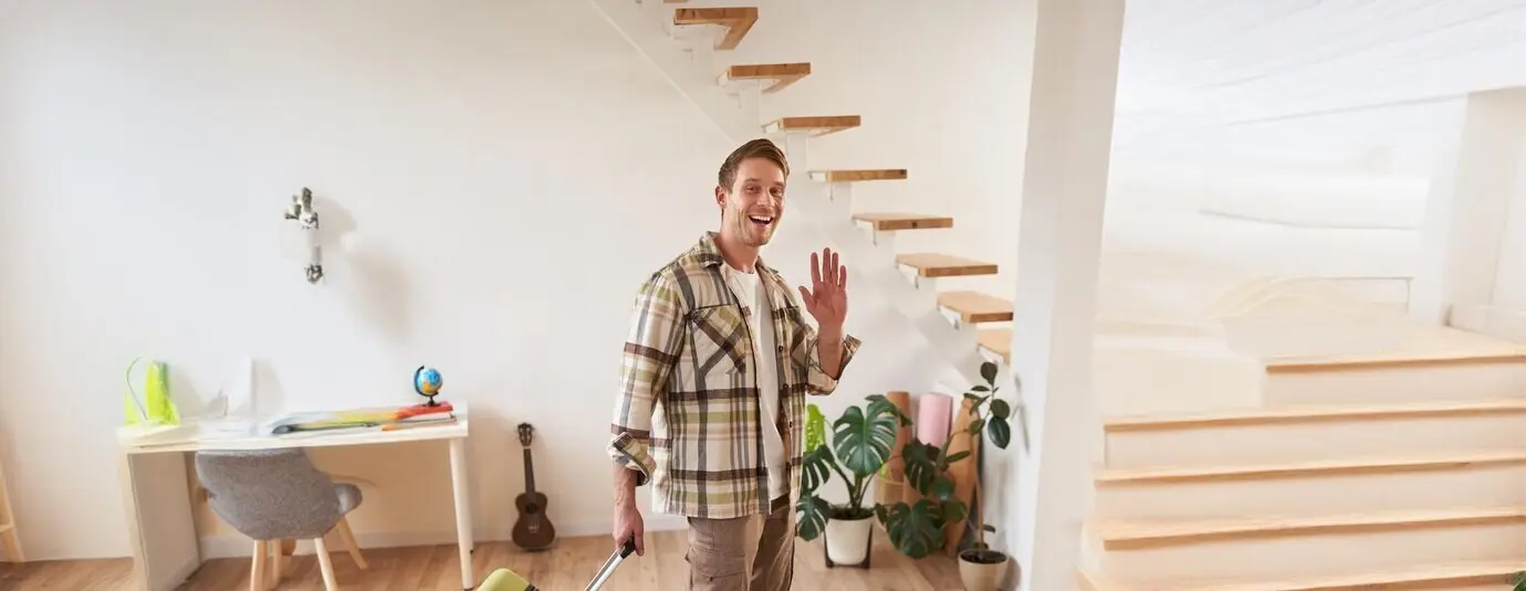 Hochformataufnahme eines fröhlichen jungen Mannes mit Koffer, der beim Einchecken in der gemieteten Wohnung der Kamera zuwinkt und drinnen mit Gepäck geht.